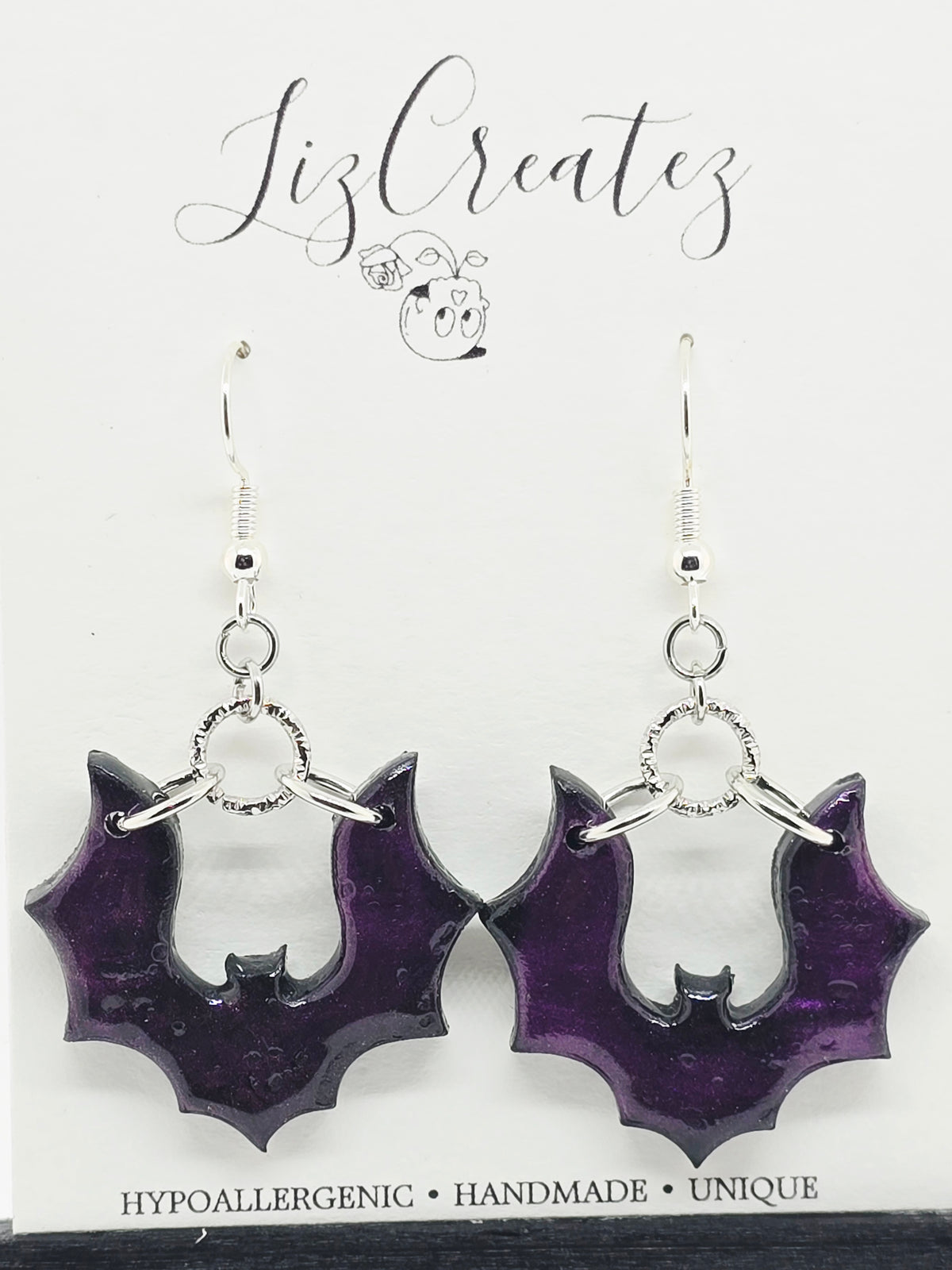 Pair of handmade purple bat-shaped polymer clay dangle earrings with glossy translucent wings, silver-tone hooks, and circular connectors, displayed against a black background. Gothic Halloween-inspired jewelry by LizCreatez.