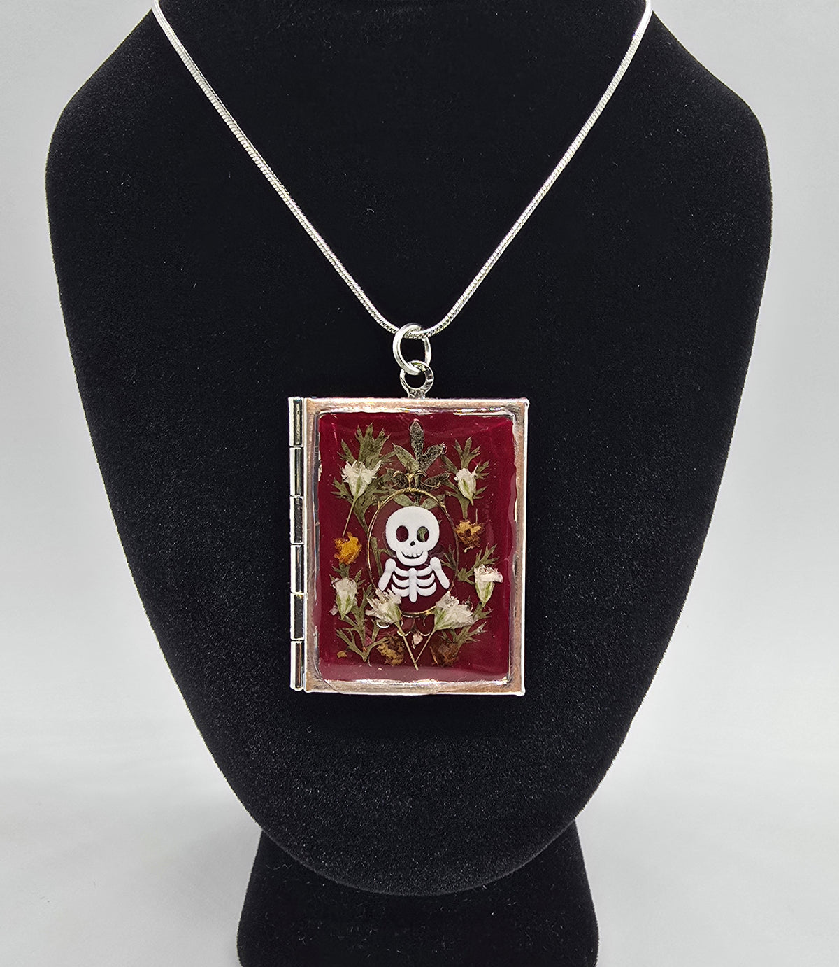 Book-shaped locket necklace with a skeleton framed in real fall leaves and flowers on burgundy, shown closed and open to reveal two oval photo frames
